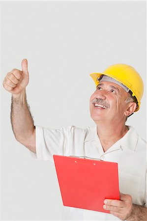 Happy senior constructor holding clipboard while gesturing thumbs up over gray background Stock Photo - Premium Royalty-Free, Code: 693-06378939