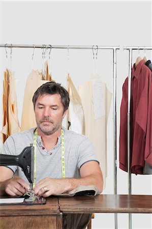 Male dressmaker stitching cloth on sewing machine with clothes rack in background Stock Photo - Premium Royalty-Free, Code: 693-06379009