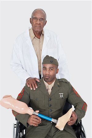 elderly not illustration not monochrome and two people assistance not illustration not monochrome and two people - Portrait of senior doctor with military officer holding artificial limb as he sits in wheelchair, studio shot on gray background Foto de stock - Sin royalties Premium, Código: 693-06379090