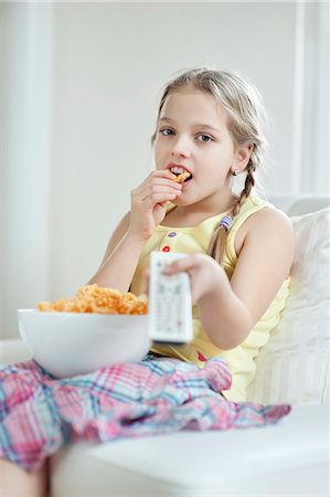 Little girl watching TV as she eats wheel shape snack pellets Stock Photo - Premium Royalty-Free, Code: 693-06379391