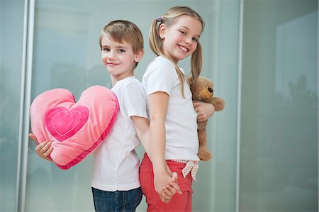 Boy and girl with heart shape cushion and teddy bear holding hands while standing back to back Foto de stock - Royalty Free Premium, Número: 693-06379440