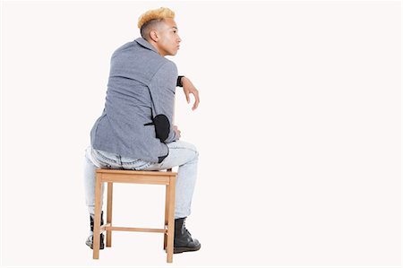 Back view of teenage boy sitting on chair as he looks away over gray background Stock Photo - Premium Royalty-Free, Code: 693-06379517