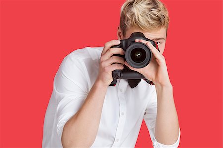 simsearch:693-06379561,k - Young man taking picture with digital camera over red background Fotografie stock - Premium Royalty-Free, Codice: 693-06379559