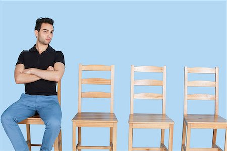 Portrait of a young man sitting beside empty wooden chairs over blue background Stock Photo - Premium Royalty-Free, Code: 693-06379590