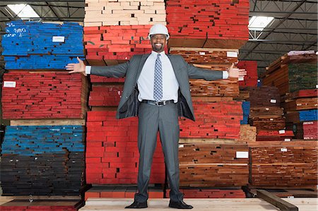 Portrait of happy African American male engineer standing with arms outstretched in front of stacked wooden planks Stock Photo - Premium Royalty-Free, Code: 693-06379742