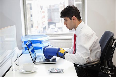 simsearch:693-06379784,k - Young Indian businessman wearing boxing gloves while using laptop at office desk Fotografie stock - Premium Royalty-Free, Codice: 693-06379781