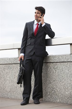 Young Indian businessman using cell phone with laptop bag by parapet Stock Photo - Premium Royalty-Free, Code: 693-06379794