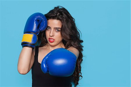 portrait of woman boxer - Portrait of an aggressive young woman wearing boxing gloves over blue background Stock Photo - Premium Royalty-Free, Code: 693-06379854