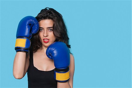 portrait of woman boxer - Portrait of an aggressive young woman wearing boxing gloves over blue background Stock Photo - Premium Royalty-Free, Code: 693-06379853