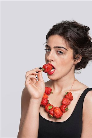 Portrait of a young woman wearing strawberry necklace as she eats one piece over gray background Stock Photo - Premium Royalty-Free, Code: 693-06379864