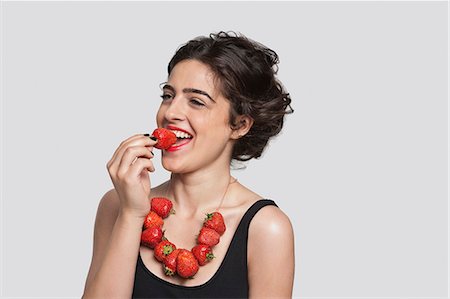 people with fruits cutout - Happy young woman wearing strawberry necklace as she eats one piece over gray background Foto de stock - Sin royalties Premium, Código: 693-06379863