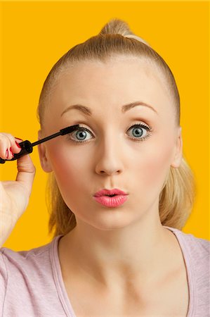 Portrait of a beautiful young woman applying mascara over yellow background Stock Photo - Premium Royalty-Free, Code: 693-06403282