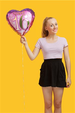 Portrait of beautiful young woman holding heart shaped birthday balloon over yellow background Stock Photo - Premium Royalty-Free, Code: 693-06403296