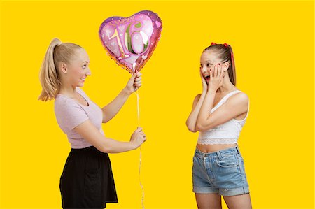 string (not clothing, packaging or instruments) - Happy young woman giving birthday balloon to amazed woman standing over yellow background Foto de stock - Sin royalties Premium, Código: 693-06403298