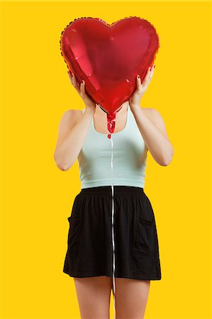 Jeune femme se cachant derrière le ballon en forme de coeur sur fond jaune Photographie de stock - Premium Libres de Droits, Code: 693-06403292