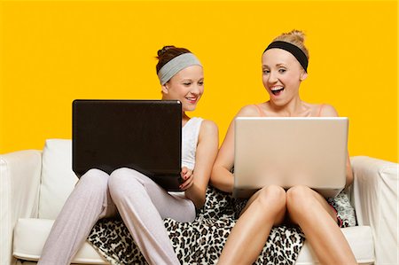 Zwei glückliche junge Frauen mit Laptop sitzen auf dem Sofa vor gelbem Hintergrund Stockbilder - Premium RF Lizenzfrei, Bildnummer: 693-06403307