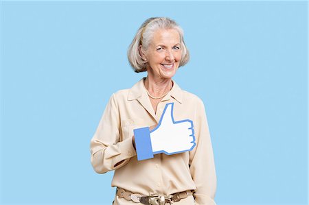 senior happy studio shot - Portrait of senior woman in casuals holding fake like button against blue background Stock Photo - Premium Royalty-Free, Code: 693-06403414