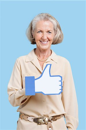 phoney - Portrait of senior woman in casuals holding fake like button against blue background Stock Photo - Premium Royalty-Free, Code: 693-06403415