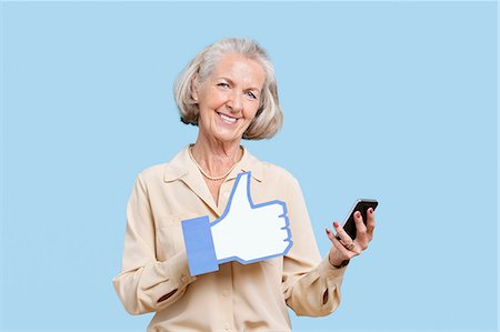 Portrait of senior woman with cell phone holding fake like button against blue background Stock Photo - Premium Royalty-Free, Code: 693-06403417