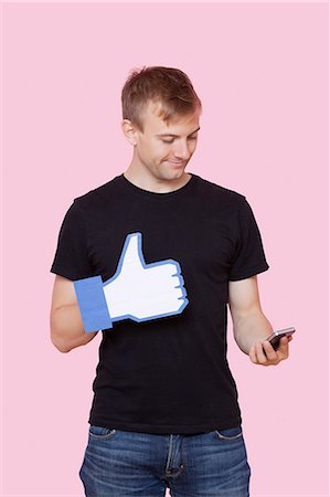 Young man with cell phone holding fake like button against pink background Stock Photo - Premium Royalty-Free, Code: 693-06403524