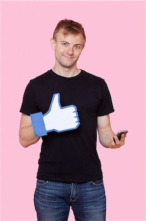 Portrait of a happy young man with cell phone holding fake like button over pink background Foto de stock - Sin royalties Premium, Código: 693-06403525