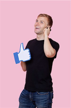 Cheerful young man using cell phone while holding fake like button over pink background Stock Photo - Premium Royalty-Free, Code: 693-06403526