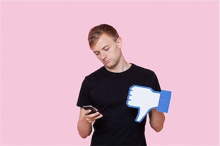 Young man with cell phone holding fake dislike button against pink background Stock Photo - Premium Royalty-Free, Code: 693-06403527