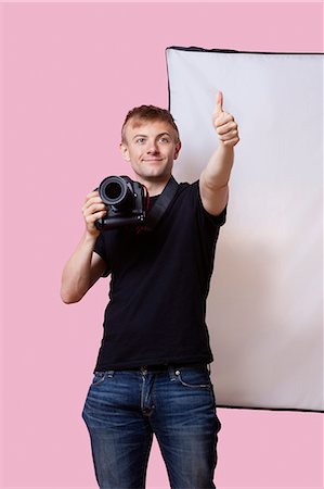 photographer - Happy photographer holding camera with thumbs up gesture over pink background Stock Photo - Premium Royalty-Free, Code: 693-06403535