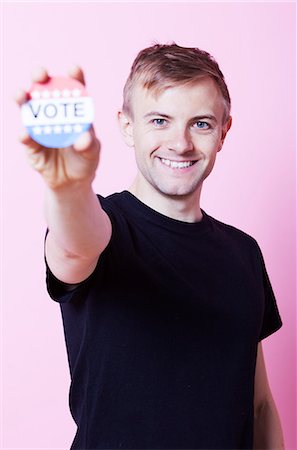 Portrait d'un jeune homme tenant un insigne VOTE jusqu'à la caméra sur fond rose Photographie de stock - Premium Libres de Droits, Code: 693-06403530