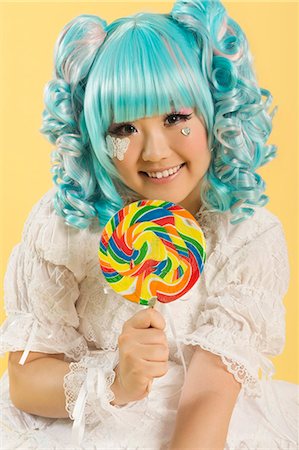 simsearch:693-06436007,k - Portrait of smiling young woman dressed as a doll holding lollipop over yellow background Stock Photo - Premium Royalty-Free, Code: 693-06436075