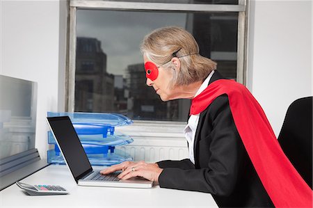 senior woman with laptop - Side view of senior businesswoman in superhero costume using laptop at office desk Stock Photo - Premium Royalty-Free, Code: 693-06497646