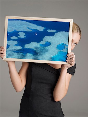 simsearch:693-06668015,k - Young businesswoman peeking from behind a photograph of cruise ship on water against gray background Photographie de stock - Premium Libres de Droits, Code: 693-06668018