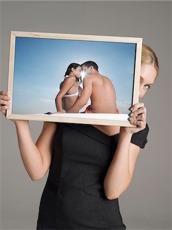 simsearch:693-06668015,k - Young businesswoman peeking from behind photograph of couple at beach against gray background Photographie de stock - Premium Libres de Droits, Code: 693-06668019