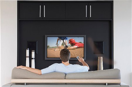 Back view of mid-adult man changing channels with television remote control in living room Stock Photo - Premium Royalty-Free, Code: 693-06668025