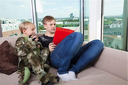 Boy dressed in dinosaur costume sitting with father reading story book on sofa bed at home Foto de stock - Sin royalties Premium, Código: 693-07455871