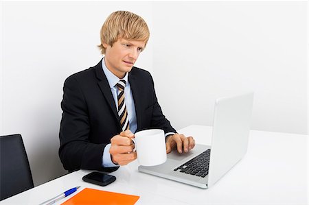 simsearch:693-06379459,k - Businessman holding coffee cup while using laptop at desk in office Stock Photo - Premium Royalty-Free, Code: 693-07455973
