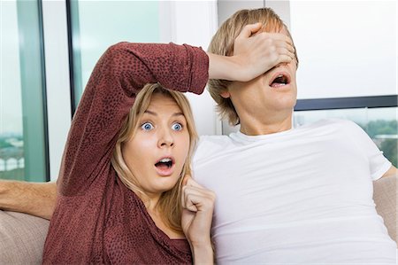 Shocked woman covering man's eyes while watching TV at home Stock Photo - Premium Royalty-Free, Code: 693-07456034