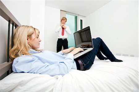 Businesswoman using laptop in bed with businessman adjusting tie at hotel Stock Photo - Premium Royalty-Free, Code: 693-07456088