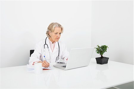 Senior female doctor writing notes while looking at laptop in clinic Stock Photo - Premium Royalty-Free, Code: 693-07456469