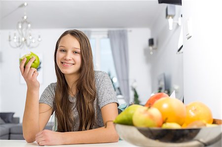 simsearch:693-07542115,k - Portrait of smiling girl holding apple while sitting at table in house Stock Photo - Premium Royalty-Free, Code: 693-07673215