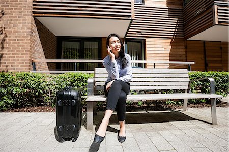 simsearch:693-06379884,k - Full length of businesswoman answering cell phone while sitting by luggage on bench against building Stock Photo - Premium Royalty-Free, Code: 693-07912095