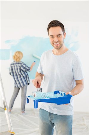 Portrait of man holding paint roller and tray with woman painting wall in background Stock Photo - Premium Royalty-Free, Code: 693-07912659