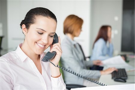 simsearch:693-07913227,k - Smiling young businesswoman using landline telephone with colleagues in background at office Foto de stock - Royalty Free Premium, Número: 693-07913217