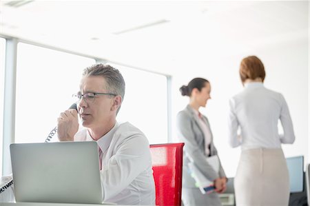 simsearch:693-07913272,k - Businessman talking on telephone with colleagues in background at office Photographie de stock - Premium Libres de Droits, Code: 693-07913249