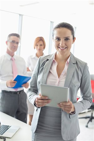 simsearch:693-07913272,k - Portrait of happy businesswoman holding digital tablet with colleagues in background at office Photographie de stock - Premium Libres de Droits, Code: 693-07913254