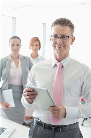 simsearch:693-07913272,k - Portrait of happy businessman holding digital tablet with female colleagues in background at office Photographie de stock - Premium Libres de Droits, Code: 693-07913253