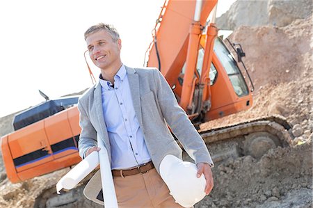 simsearch:693-08127789,k - Smiling architect looking away while holding blueprints and hardhat at construction site Stock Photo - Premium Royalty-Free, Code: 693-08127793