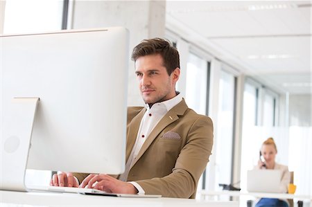 simsearch:693-08769254,k - Confident young businessman working on computer with female colleague in background at office Foto de stock - Sin royalties Premium, Código: 693-08769266