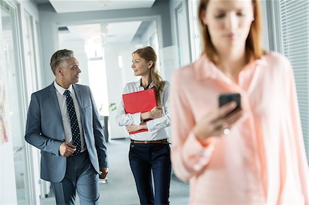 simsearch:693-08769289,k - Business people walking in corridor with female colleague using mobile phone in foreground at office Stock Photo - Premium Royalty-Free, Code: 693-08769314