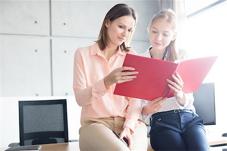 simsearch:6109-06005438,k - Young businesswomen reading file while sitting on desk in office Photographie de stock - Premium Libres de Droits, Code: 693-08769334